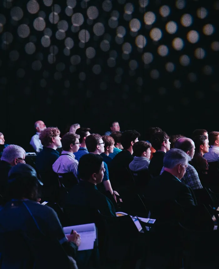 Audience seated in a dark conference hall, some taking notes